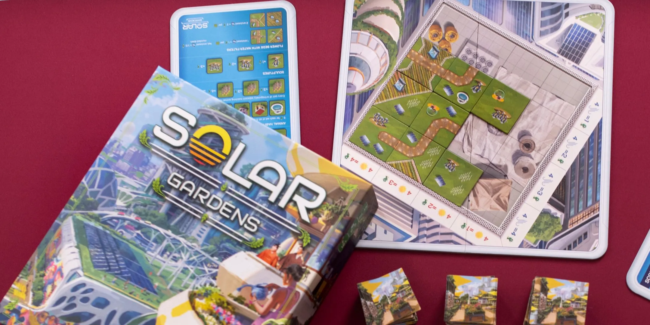 Tổng quan hộp và các thành phần của board game Solar Gardens được sắp xếp trên nền đỏ.